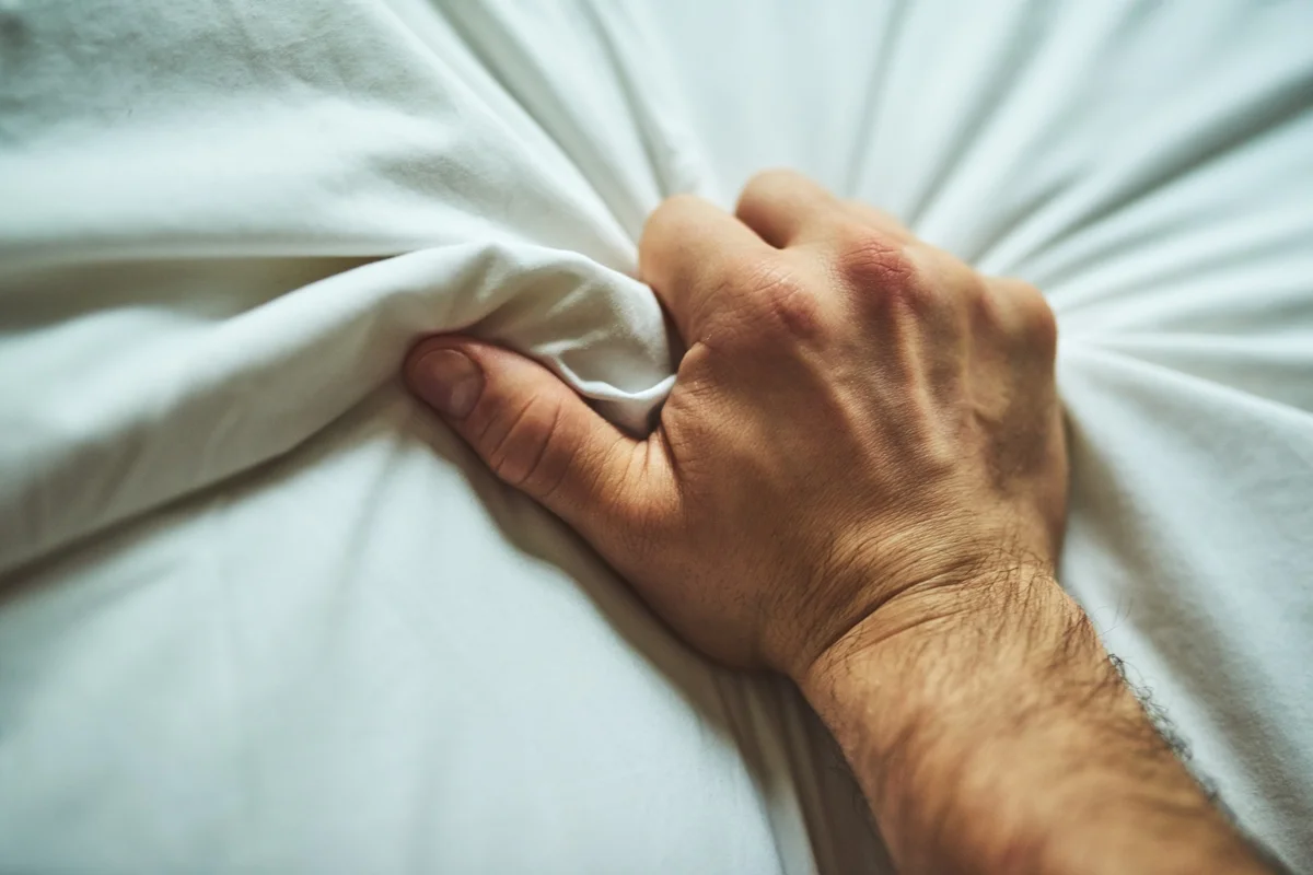 Foto em close da mão de um homem irreconhecível agarrando um lençol com prazer durante masturbação